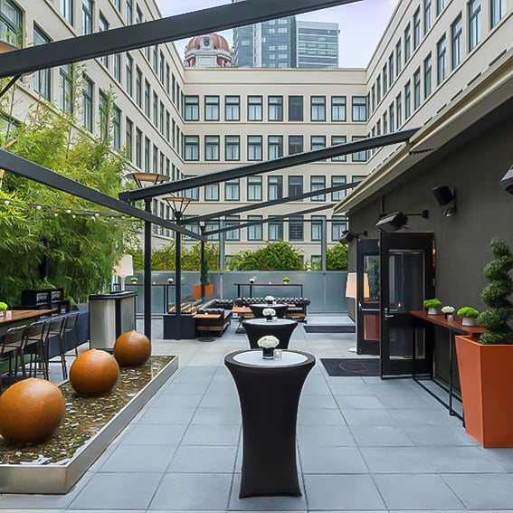 A stylish, modern outdoor courtyard set up for an event, featuring high-top cocktail tables with white floral centerpieces, sleek seating areas, water features with decorative spheres, and city buildings in the background. The space is inviting and ready for guests to mingle.