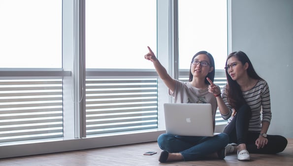 Two women sitting cross legged with a laptop in front of windows, pointing at something in the distance.