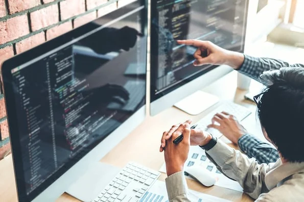 two people sitting at a desk using a desktop computer to edit, customize, and override Bootstrap CSS