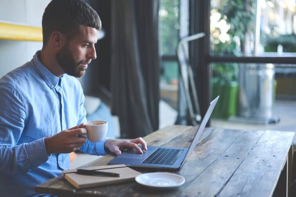 Mann hält Kaffeetasse in einer Hand und arbeitet an einem Laptop