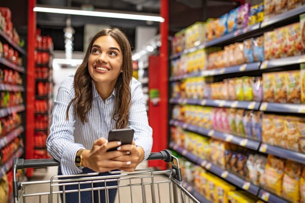 Frau mit Einkaufswagen kauft im Supermarkt ein und hält ihr Smartphone in den Händen 