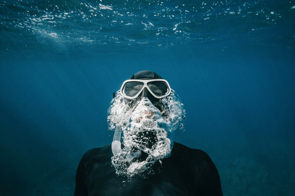 Mann mit Taucherbrille stößt Luftblasen im Wasser aus