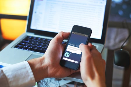 Hand hält Smartphone mit Chatbot vor Laptop