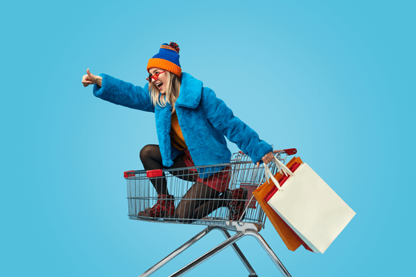 Frau kniet in Einkaufswagen mit Einkaufstüten in der Hand