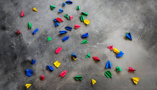 Paper airplanes in Google colors of red, green, blue, and yellow scattered on gray pavement.