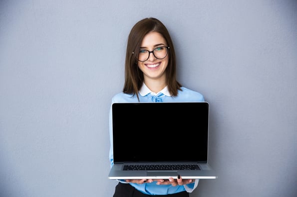 Happy businesswoman showing blank laptop screen over gray background. Wearing in blue shirt and glasses. Looking at camera.jpeg