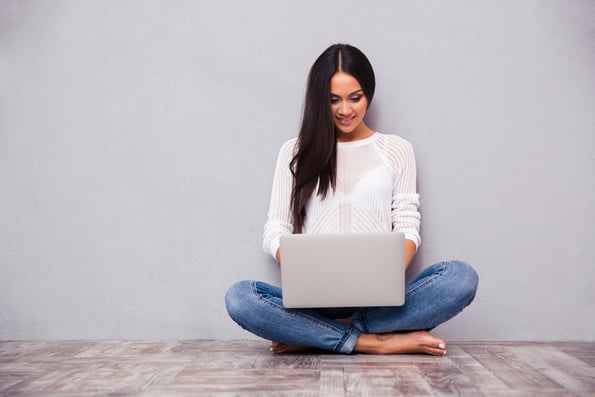 Portrait of a happy casual woman sitting on the floor with laptop on gray background.jpeg