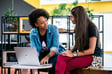 Two women engaged in a conversation, sitting at a table with a laptop open, surrounded by indoor plants and a modern workspace background.