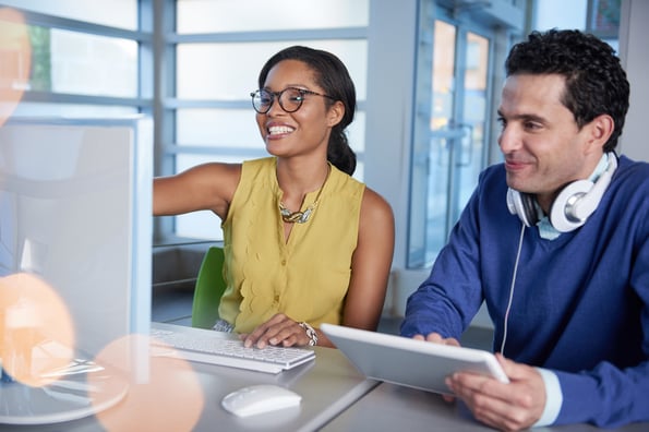 Coworkers looking at a computer screen. The woman is pointing at it and the man is holding a tablet in his hands.