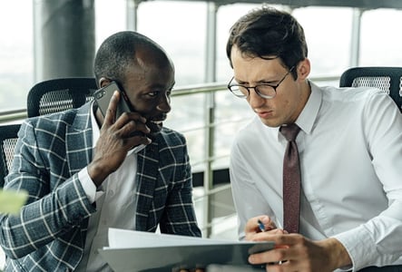 Two businessmen analyze a document. One is on the phone and the other is pointing at the paper with a pen.