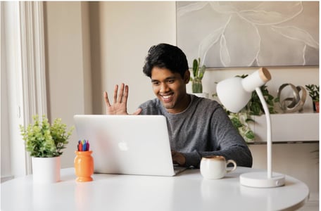 Vendedor conversa com cliente em ambiente de trabalho remoto - vendedor está sentado a frente de um notebook e acena para cliente em chamada de vídeo 