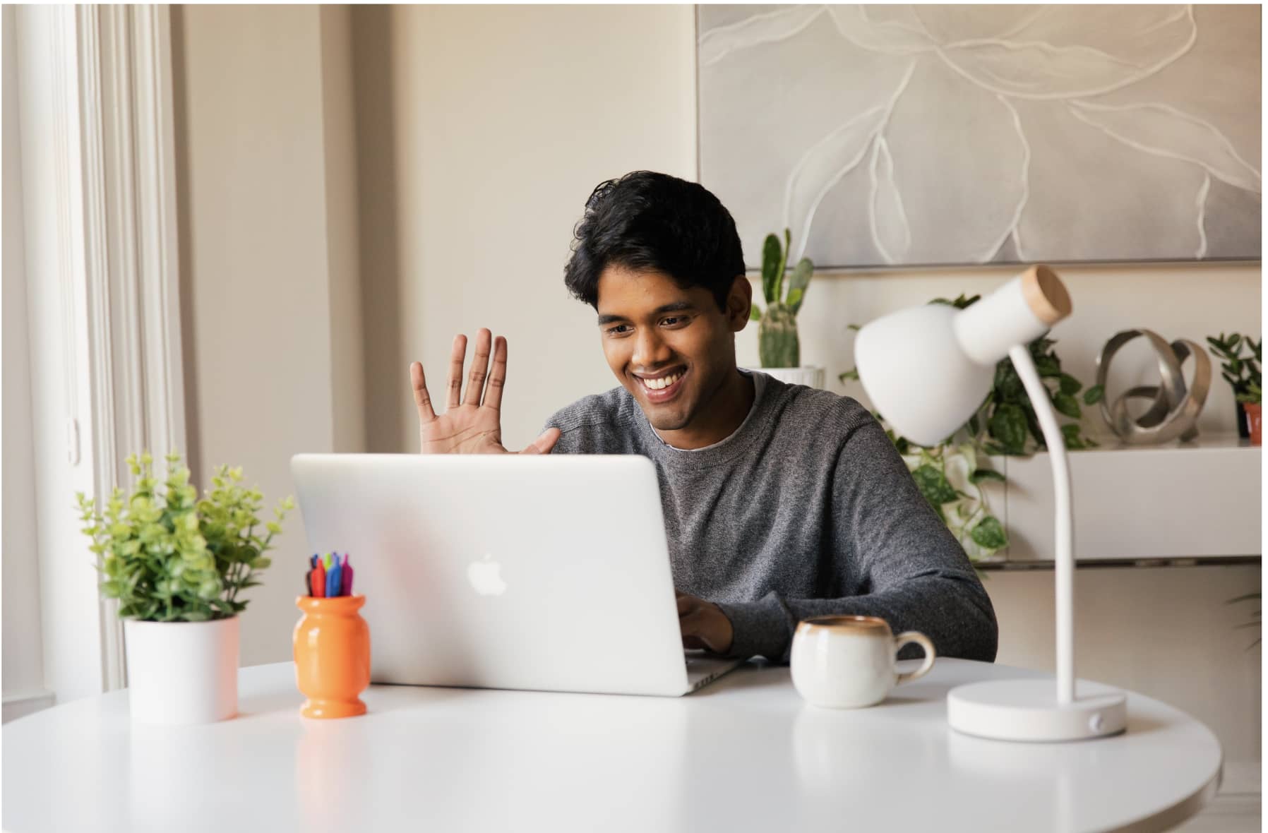 Vendedor conversa com cliente em ambiente de trabalho remoto - vendedor está sentado a frente de um notebook e acena para cliente em chamada de vídeo 